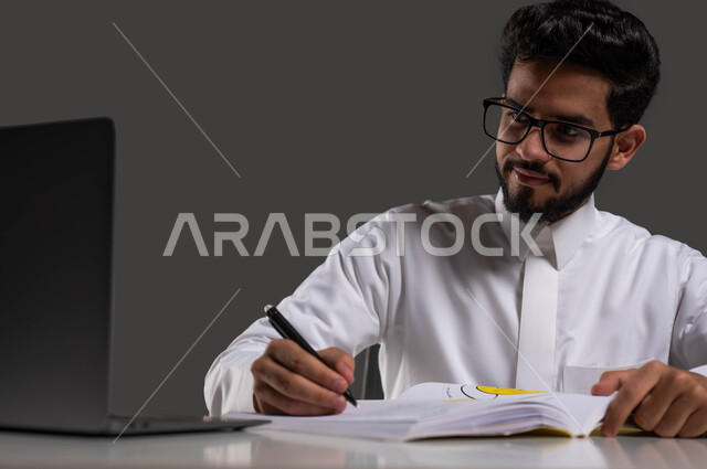 Following lessons and assignments online, developing educational curricula in Saudi Arabia, the importance of education in developing Saudi society, a close-up portrait of a Saudi high school student wearing traditional clothing and glasses, sitting in front of the study table and computer and writing his homework, black background.