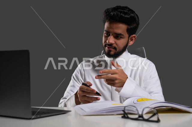 تفاعل الطلاب مع المحتوى التعليمي عبر الانترنت، المقررات والمناهج التعليمية الحديثة والمطورة بالسعودية، بورتريه لطالب جامعي عربي خليجي سعودي يرتدي الزي التقليدي يجلس أمام الطاولة الدراسية وجهاز الحاسوب، خلفية سوداء.