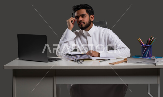 المقررات والمناهج التعليمية الحديثة والمطورة بالسعودية، دور المدارس في رفع مستوى الوعي لدى الطلاب، بورتريه لطالب ثانوي عربي خليجي سعودي يرتدي الزي التقليدي يجلس أمام الطاولة الدراسية بإيماءات وجه ويدين تدل على التفكير والشرود، خلفية سوداء.