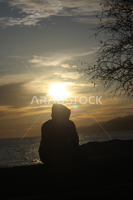 التأمل والنظر في مياه البحر الهادئة، صورة ظلية من الخلف لشاب يجلس على شاطئ البحر، الطبيعة البحرية وقت غروب الشمس، أماكن سياحية ترفيهية، سيلوليت