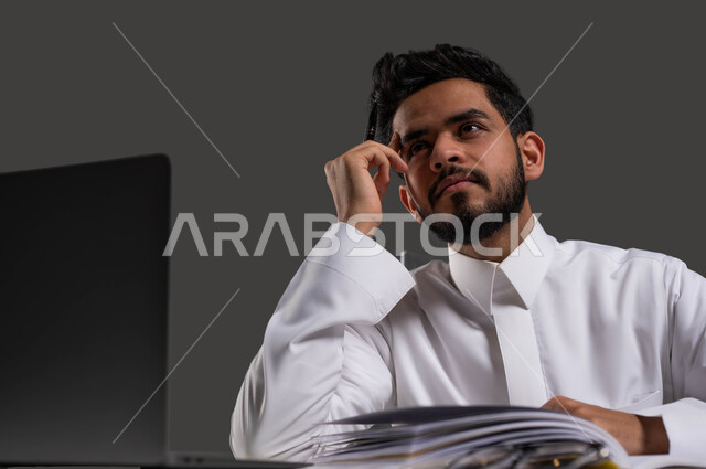 The importance of education in building a bright future for Saudi society, the role of schools in raising the level of awareness among students, a close-up portrait of a Saudi Arabian Gulf high school student wearing traditional clothing sitting in front of the study table with facial and hand gestures indicating thought and distraction, a black background.