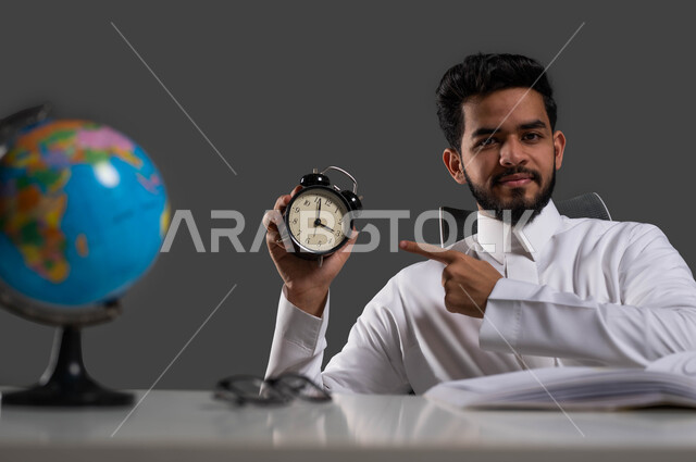 استغلال الوقت في الدراسة والمذاكرة، تطوير المناهج التعليمية في السعودية، بورتريه لطالب ثانوي عربي خليجي سعودي يرتدي الثوب التقليدي يجلس على طاولة الدراسة ينظر إلى الكاميرا ويشير بإصبع السبابة إلى ساعة المنبه, خلفية سوداء.