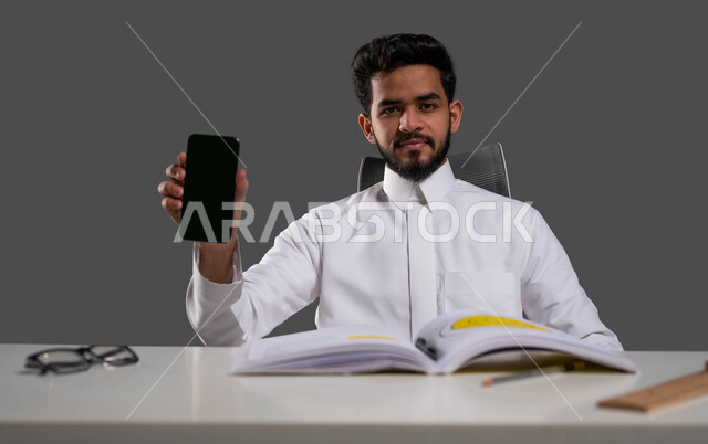 استخدام الأجهزة الذكية والتطبيقات في التعلم عن بعد، دور التكنولوجيا في تحسين أداء الطلاب، بورتريه مقرب لطالب جامعي عربي خليجي سعودي يرتدي الزي التقليدي يجلس على المكتب الدراسي ويمسك بيده الهاتف المحمول بشاشة سوداء فارغة، النظر إلى الكاميرا، خلفية سوداء.