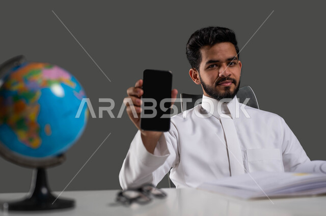 مراجعة الدروس اليومية عبر الانترنت، استخدام الأجهزة الذكية والتطبيقات في التعلم عن بعد، بورتريه مقرب لطالب جامعي عربي خليجي سعودي يرتدي الزي التقليدي يجلس على الطاولة ويمسك بيده الهاتف المحمول بشاشة سوداء فارغة، النظر إلى الكاميرا، خلفية سوداء.