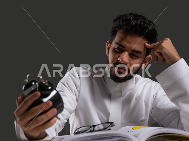 استغلال الوقت في المذاكرة والدراسة لساعات طويلة، تطوير المناهج التعليمية في السعودية، بورتريه لطالب ثانوي عربي خليجي سعودي يرتدي الثوب التقليدي يجلس على طاولة الدراسة يمسك بيده ساعة المنبه بإيماءات التفكير والشرود الذهني, خلفية سوداء.