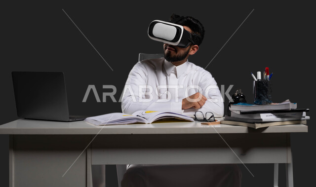 جودة التعليم المتطور باستخدام تقنية العالم الافتراضي في السعودية، دور نظارات الواقع الافتراضي في تعزيز التفاعل والمشاركة والانسجام في الفصول الدراسية، بورتريه لطالب ثانوي عربي خليجي سعودي يرتدي الزي التقليدي وبجلس على الطاولة، خلفية سوداء.