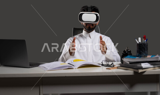 جودة التعليم المتطور باستخدام تقنية العالم الافتراضي في السعودية، دور نظارات الواقع الافتراضي في تعزيز التفاعل والمشاركة في الفصول الدراسية، بورتريه لطالب جامعي سعودي يرتدي الزي التقليدي ويشير بيديه إلى الأمام بإيماءات التحجيم والإمساك بعنصر ما، خلفية سوداء.