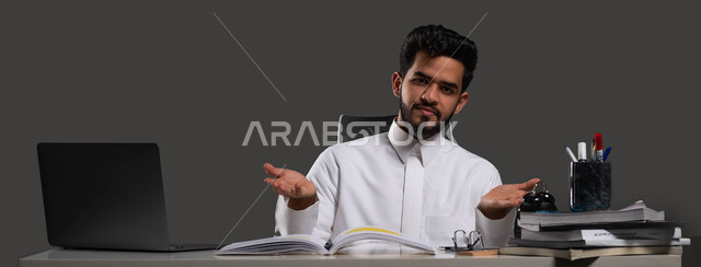 الصعوبة في فهم الدرووس والمسائل، النظر إلى الكاميرا بإيماءات الحيرة وعدم المعرفة، الحاجة إلى المساعدة في المذاكرة، بورتريه مقرب لطالب جامعي عربي خليجي سعودي يرتدي الزي التقليدي يجلس على المكتب الدراسي، خلفية سوداء.