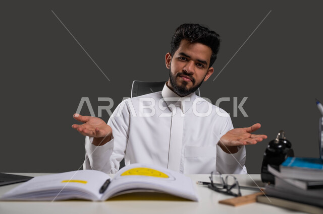 النظر إلى الكاميرا بإيماءات الحيرة وعدم المعرفة، الصعوبة في فهم الدرووس والمسائل، الحاجة إلى المساعدة في المذاكرة، بورتريه مقرب لطالب جامعي عربي خليجي سعودي يرتدي الزي التقليدي يجلس على المكتب الدراسي، خلفية سوداء.