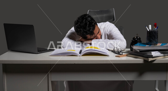 دخول وقت النوم، المذاكرة لأوقات متاخرة من الليل، التعب والاجهاد اثناء مراجعة الدروس اليومية، السعي للتفوق والنجاح، بورتريه لطالب ثانوي عربي خليجي سعودي يرتدي الزي التقليدي نائم على طاولة الدراسة، خلفية سوداء.