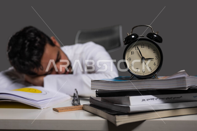 Fatigue and stress while reviewing daily lessons, studying late into the night, striving for excellence and success, close-up portrait of a Saudi Gulf Arab high school student wearing traditional clothing sleeping at the study table, entering bedtime, black background.