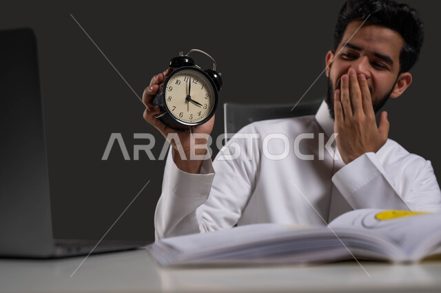 The difficulty of waking up early for school, the end of study time and the beginning of bedtime, a close-up portrait of a Saudi Arabian Gulf university student wearing traditional clothing, holding an alarm clock in his hand and placing his hand over his mouth with gestures indicating fatigue and sleepiness, black background.