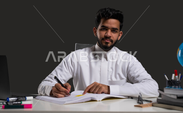 تطوير المناهج والمواد التعليمية في السعوديه، حل الواجبات وكتابة الملاحظات، أهمية التعليم في تطوير المجتمع السعودي، بورتريه مقرب لطالب ثانوي عربي خليجي سعودي يرتدي الزي التقليدي وينظر إلى الكاميرا، خلفية سوداء.