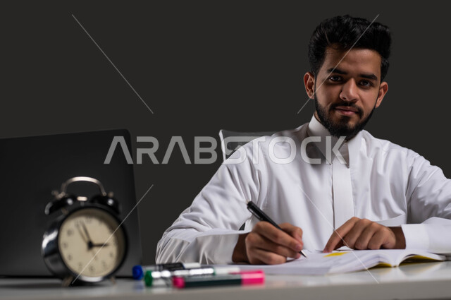 Doing assignments and writing notes, developing curricula and educational materials in Saudi Arabia, the importance of education in developing Saudi society, a close-up portrait of a Saudi Gulf Arab high school student wearing traditional clothing and looking at the camera, black background.