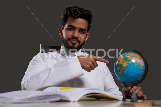 الاهتمام بدراسة العلوم الجغرافية، توسيع الآفاق وفهم التنوع الثقافي والطبيعي، أهمية التعليم في تطوير المجتمع السعودي، بورتريه مقرب لطالب جامعي عربي خليجي سعودي يرتدي الزي التقليدي ويشير بيده الى مجسم الكرة الأرضية، خلفية سوداء.