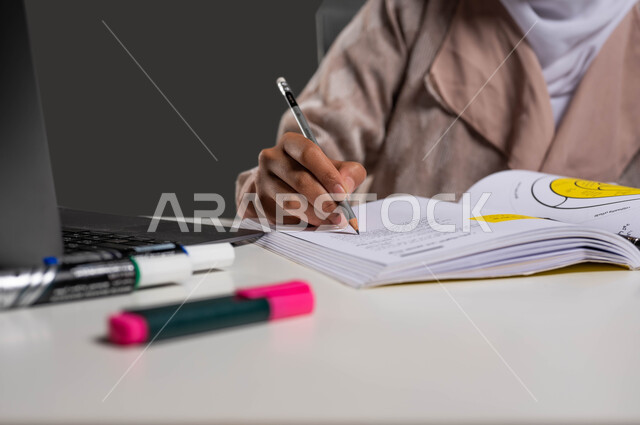 دور التكنولوجيا في تغيير أساليب التعليم التقليدية, مفهوم التعليم عبر جهاز الحاسوب, بورتريه مقرب لطالبة جامعية عربية خليجية سعودية تجلس على طاولة الدراسة وتقوم بكتابة واجباتها المدرسية, خلفية سوداء.