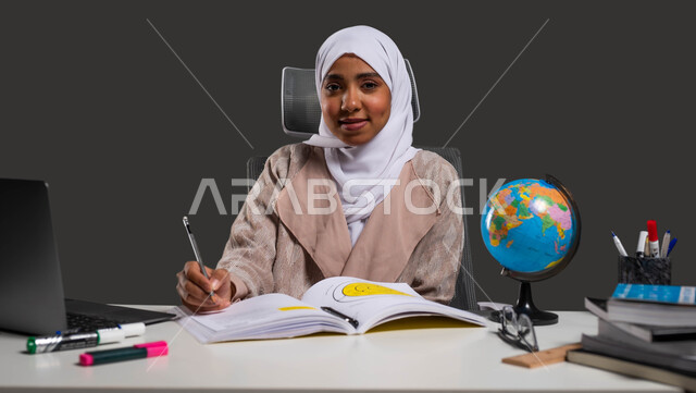 Using a computer to solve school assignments, the concept of online education, a portrait of a veiled Saudi Gulf Arab high school student wearing an abaya, sitting at the study table and looking at the camera, developing students’ skills through virtual education, black background.