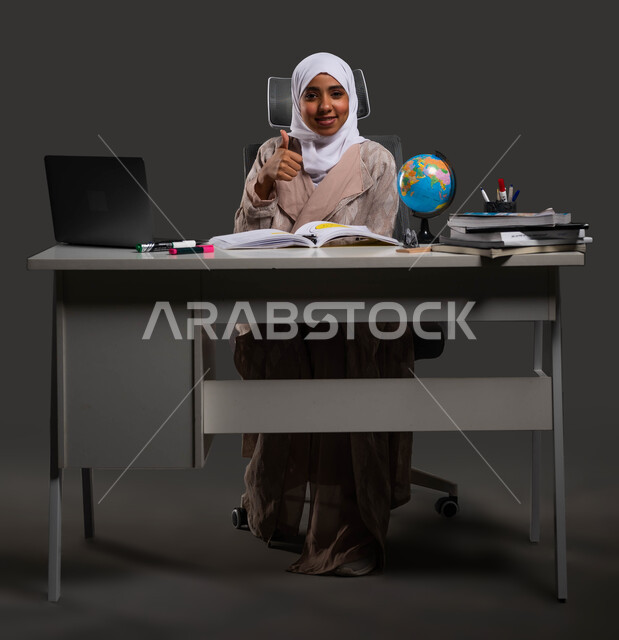 The concept of home education via a laptop, a portrait of a veiled Saudi Gulf Arab high school student wearing an abaya and raising her thumb up, the role of technology in changing traditional teaching methods, gestures of acceptance and approval, a black background.