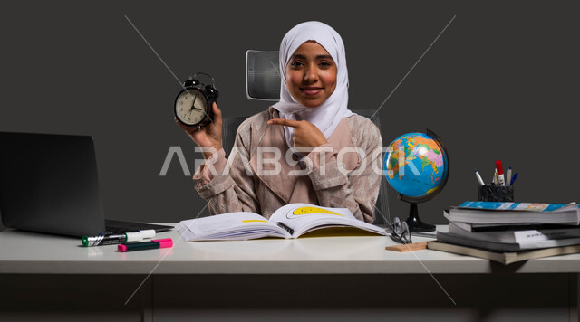 The arrival of exams, using time to study and study, developing educational curricula in Saudi Arabia, spending enjoyable times in home education, a portrait of a veiled Saudi high school student wearing an abaya, looking at the camera and pointing to an alarm clock, black background.