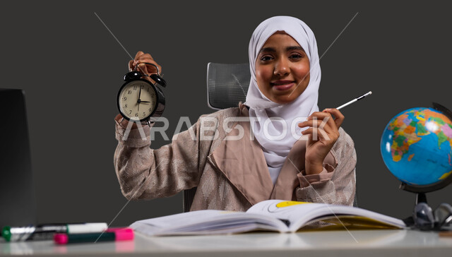 قضاء وقت ممتع في التعليم المنزلي، النظر إلى الكاميرا بإيماءات تدل على السرور، استغلال الوقت في الدراسة والمذاكرة, بورتريه مقرب لطالبة جامعية عربية خليجية سعودية محجبة ترتدي العباءة وتحمل بيدها ساعة منبه, خلفية سوداء.
