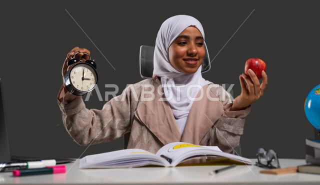 أهمية الطعام الصحي في زيادة تركيز الطلاب، استغلال الوقت في الدراسة والمذاكرة، بورتريه مقرب لطالبة ثانوية عربية خليجية سعودية محجبة ترتدي العباءة وتحمل بيديها ساعة منبه وتفاحة حمراء, إيماءات الفرح بدخول وقت الفسحة، خلفية سوداء.