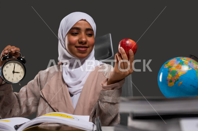 تحسين مستوى الذكاء لدى الطلاب بتناول الغذاء الصحي، استغلال الوقت في الدراسة والمذاكرة، بورتريه مقرب لطالبة ثانوية عربية خليجية سعودية محجبة ترتدي العباءة وتحمل بيديها ساعة منبه وتفاحة حمراء, إيماءات الفرح بدخول وقت الفسحة، خلفية سوداء.