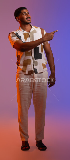 التأمل وتحديد الأهداف، حركات تدل على الذهاب إلى مكان ما، بورتريه لرجل عربي خليجي سعودي مبتسم يرتدي الزي الكاجوال يشير بأصابعه نحو اليمين بإيماءات السرور، مفهوم لغة الجسد وتعابير الوجه، صورة الجسم بالطول الكامل، خلفية ملونة