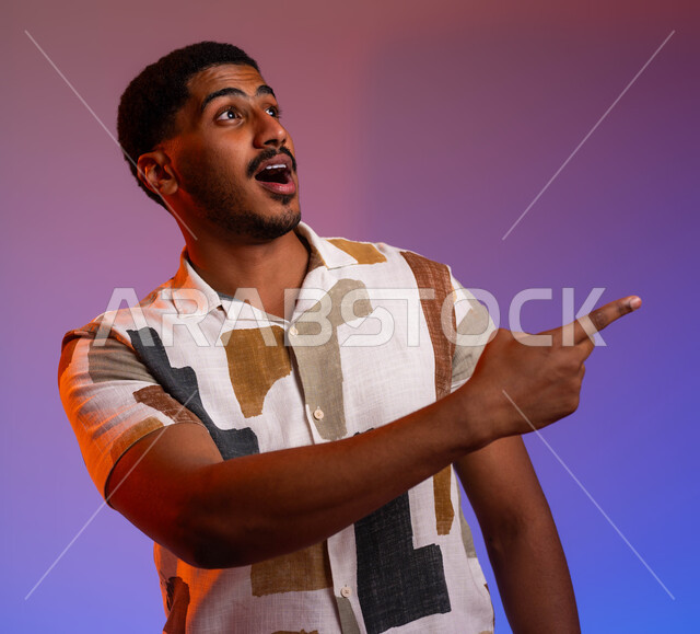 الاستغراب من أمر معين، الشعور بالصدمة والذهول، حركات ودلالات مختلفة، بورتريه مقرب لرجل عربي خليجي سعودي يرتدي الزي الكاجوال يشير بأصابعه نحو اليمين بإيماءات التعجب والاندهاش من شيء ما، خلفية ملونة