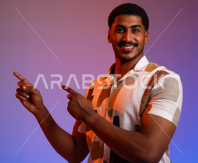 حركات ودلالات مختلفة، إيماءات السعاده و السرور في تحديد موقع معين، بورتريه مقرب لشاب عربي خليجي سعودي مبتسم يرتدي الزي الكاجوال يشير بأصابعه نحو الأعلى وينظر إلى الكاميرا،  خلفية ملونة
