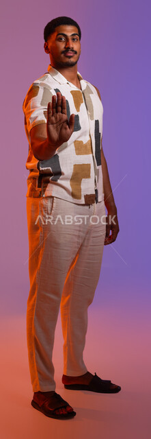 الشعور بالغرور والثقة بالنفس، إيماءات تدل على التشكر وعدم الحاجة للمساعدة, طلب الصمت والتوقف عن الكلام، بورتريه مقرب لرجل عربي سعودي خليجي يرتدي الزي الكاجوال يرفع كفه مفتوحة للأمام, صورة الجسم بالطول الكامل, خلفية ملونة