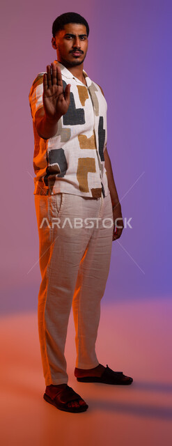 Condemning gestures indicating the command to stop doing something and forbidding it, silence and stopping talking, portrait of a Saudi Gulf Arab young man wearing casual clothes and raising his palm up, feeling of annoyance, rejection and abstinence, full-length body portrait, colored background