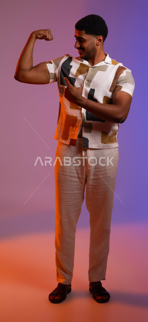 Bodybuilding, physical fitness for players, building an athletic body, portrait of a Saudi Gulf Arab young man wearing casual summer clothes, portrait of a Saudi Gulf Arab young man wearing casual summer clothes pointing with his finger to the muscle of his other hand, full body photo, colored background