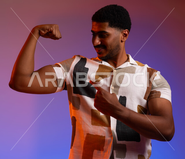 استعراض القوة البدنية، عرض عضلات اليدين،  نشاط بدني للمحافظة على صحة الجسم، بناء العضلات وتقويتها، بورتريه مقرب لشاب عربي سعودي خليجي يرتدي ملابس كاجول صيفية يرفع يده بكفة مقبوضة، خلفية ملونة.