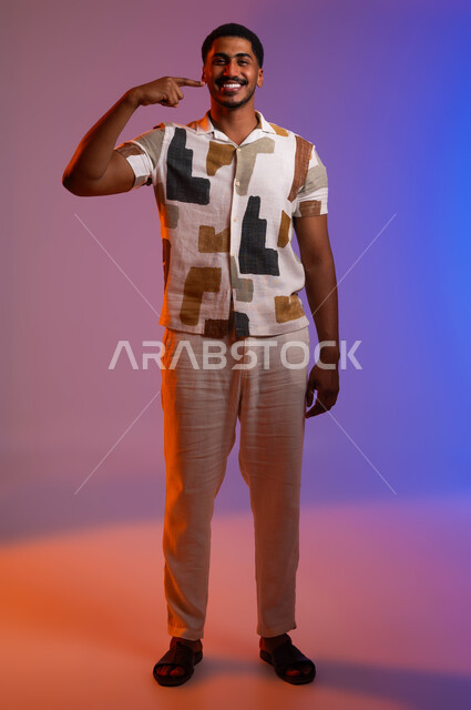 The concept of care and concern for health and appearance, beautification and whitening of teeth, portrait of a Saudi Gulf Arab man wearing a casual outfit pointing with his index finger on his white teeth, using porcelain veneers for teeth, the beauty of a Hollywood smile, full-length body portrait, white background