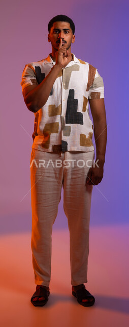 ايماءات الوجه واليدين تدل على طلب السكوت والصمت، بورتريه لرجل عربي خليجي سعودي يرتدي الزي الكاجوال يضع اصبع السبابة على فمه، التوقف عن الكلام، طلب خفض الصوت، الأمر بعدم الازعاج، صورة لكامل الجسم، خلفية ملونة