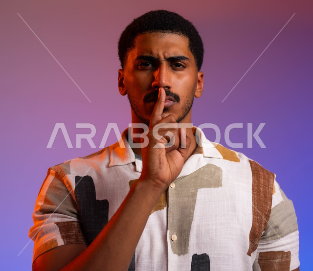 الأمر بعدم الازعاج، ايماءات الوجه واليدين تدل على طلب السكوت والصمت، بورتريه مقرب لرجل عربي خليجي سعودي يرتدي الزي الكاجوال يضع اصبع السبابة على فمه، التوقف عن الكلام، طلب خفض الصوت،  خلفية ملونة