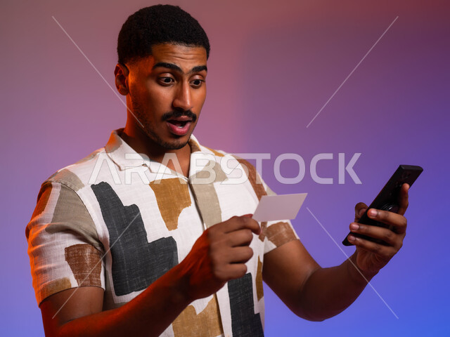 إمكانية الشراء والدفع من خلال برامج وتطبيقات عبر الجوال، ايماءات الدهشة والتعجب، بورتريه مقرب لرجل عربي سعودي خليجي يرتدي الزي الكاجوال ويمسك بيده الهاتف المحمول وبطاقة بيضاء فارغة، استخدام بطاقة الإئتمان البنكية في التسوق الالكتروني، خلفية ملونة
