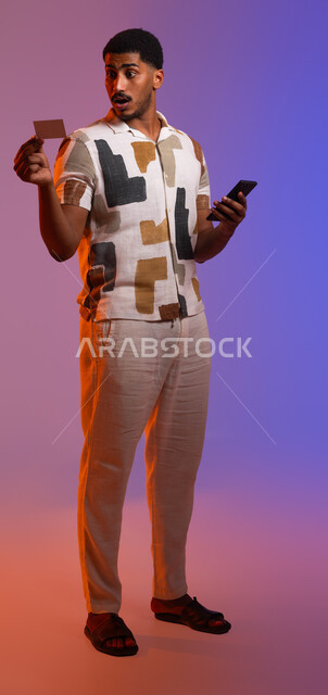 استخدام بطاقة الإئتمان البنكية في التسوق الالكتروني، إمكانية الشراء والدفع من خلال برامج وتطبيقات الجوال، ايماءات الدهشة والتعجب، بورتريه لشاب عربي سعودي خليجي يرتدي الزي الكاجوال يمسك بيده الهاتف المحمول وبطاقة بيضاء، صورة الجسم بالطول الكامل، خلفية ملونة