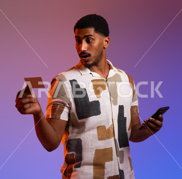 إمكانية الشراء والدفع من خلال برامج وتطبيقات عبر الجوال، ايماءات الدهشة والتعجب، بورتريه مقرب لرجل عربي سعودي خليجي يرتدي الزي الكاجوال ويمسك بيده الهاتف المحمول وبطاقة بيضاء فارغة، استخدام بطاقة الإئتمان البنكية في التسوق الالكتروني، خلفية ملونة