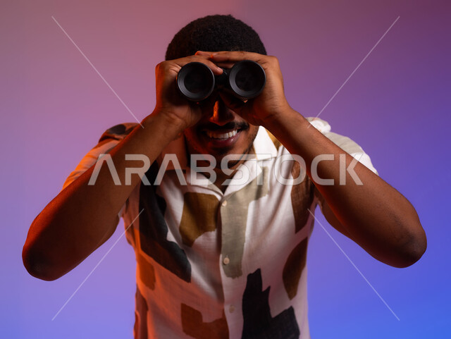 رؤية واضحة ومشرقة للمشاهد والتفاصيل، بورتريه مقرب لشاب عربي سعودي خليجي مبتسم يرتدي قميص صيفي يضع التلسكوب ثنائي العدسات على عينيه، البحث عن شيء ما، توسيع مجال الرؤية من خلال المنظار، التخطيط المستقبلي للأهداف والطموحات، خلفية ملونة