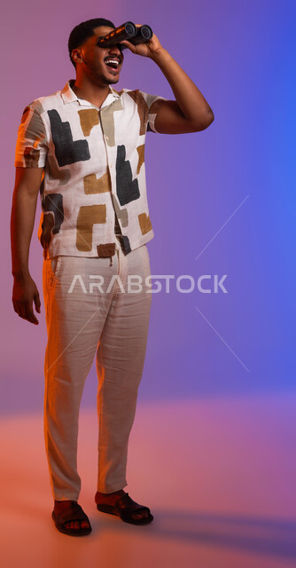 رؤية الأشياء البعيدة، بورتريه لرجل عربي سعودي خليجي مبتسم يرتدي ملابس كاجوال صيفية ويمسك بيده المنظار ثنائي العدسات، ايماءات السعادة بإيجاد شيء ما، حب الاستكشاف والمغامرة، صورة الجسم بالطول الكامل، خلفية ملونة