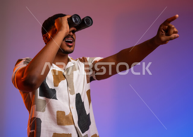 متعة الاستكشاف والمغامرة، بورتريه مقرب لشاب عربي سعودي خليجي مبتسم يرتدي ملابس كاجوال صيفية ويمسك بيده التلسكوب ثنائي العينين، توسيع مجال الرؤية من خلال المنظار، الاشارة الى شيء ما، البحث والتخطيط والنظر للمستقبل، خلفية ملونة
