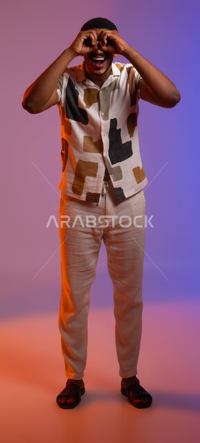 التركيز على شيء ما، بورتريه لرجل عربي سعودي خليجي مبتسم يرتدي ملابس كاجوال صيفية ويضع يده على عينه كالمنظار، ايماءات الحماس بالبحث عن شيء ما، حب الاستكشاف والمغامرة، صورة الجسم بالطول الكامل، خلفية ملونة