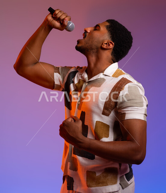 الغناء بحماس وصوت عالي، الفن المسرحي الغنائي، تقديم العروضات المسرحية، ممارسة هواية الغناء للتسلية والترفيه، بورتريه مقرب لشاب عربي سعودي خليجي مبتسم يرتدي الزي الكاجوال يحمل بيده الميكروفون، مواهب فنية في عالم الموسيقى، خلفية ملونة