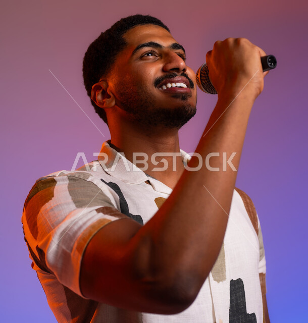 تقديم العروضات المسرحية، القاء الخطابات والحوارات والنقاشات، الفن المسرحي الغنائي، ممارسة هواية الغناء للتسلية والترفيه، بورتريه مقرب لشاب عربي سعودي خليجي مبتسم يرتدي الزي الكاجوال يحمل بيده الميكروفون، مواهب فنية في عالم الموسيقى، خلفية ملونة