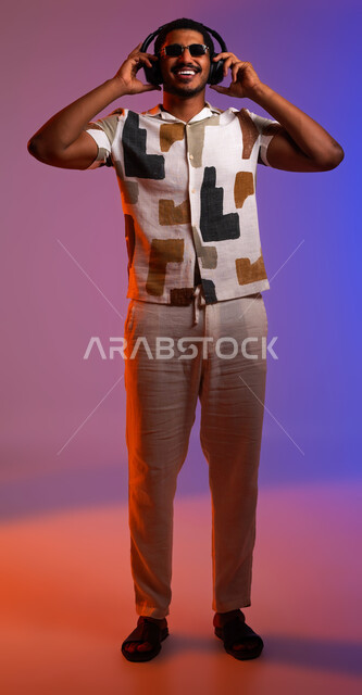 قضاء أوقات ممتعة بالإستماع للمواهب الغنائية، مفهوم الإصغاء والاستمتاع بإستخدام الهيدفون، بورتريه لرجل سعودي يرتدي الزي الكاجوال والنظارات الشمسية يضع سماعة الرأس، إيماءات وجه تدل على الاندماج والطرب، صورة الجسم بالطول الكامل، خلفية ملونة