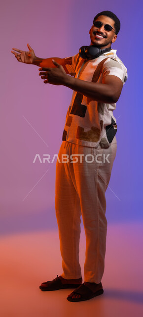 Interactions and hand movements indicating dancing and interaction with music, gestures of enthusiasm and integration, portrait of a young Saudi Arab Gulf man wearing casual clothes and sunglasses with headphones around his neck, feelings of happiness and joy, the concept of singing and rapture, full-length body portrait, colored background.