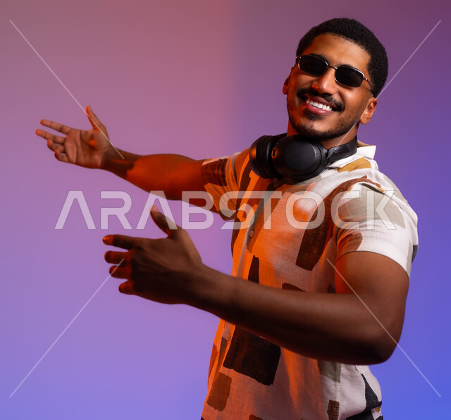 مشاعر الحب والغرام، حركات يدين تدل على الرقص والتفاعل مع الموسيقى، بورتريه مقرب لشاب عربي سعودي خليجي يرتدي الزي الكاجوال ونظارة شمسية يضع سماعات الهيدفون حول رقبته، قضاء أوقات ممتعة خارج البلاد، الغناء والطرب اثناء الرحلات السياحية، خلفية ملونة
