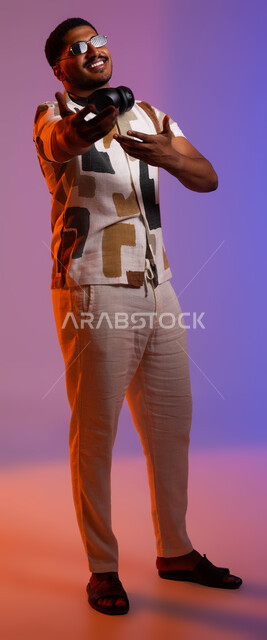 حركات يدين تدل على الرقص والتفاعل مع الموسيقى، ايماءات الحماس والاندماج، بورتريه لشاب عربي سعودي خليجي يرتدي الزي الكاجوال الصيفي والنظارات الشمسية يضع سماعات الهيدفون حول رقبته، مشاعر السعادة والفرح، مفهوم الغناء والطرب، صورة الجسم بالطول الكامل، خلفية ملونة