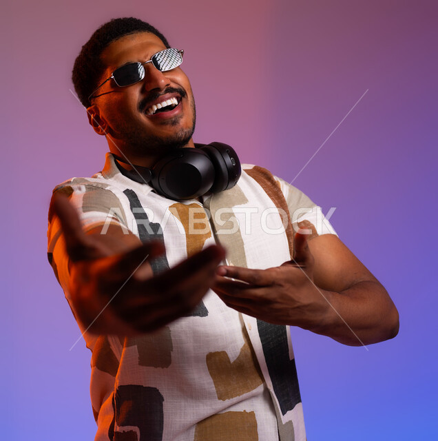 ايماءات الحماس والاندماج، بورتريه مقرب لشاب عربي سعودي خليجي يرتدي الزي الكاجوال الصيفي والنظارات الشمسية يضع سماعات الهيدفون حول رقبته، مشاعر السعادة والفرح، حركات يدين تدل على الرقص والتفاعل مع الموسيقى، مفهوم الغناء والطرب، خلفية ملونة